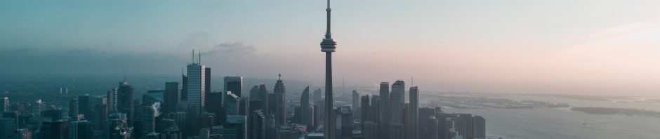 An aerial photo of downtown Toronto, including the iconic CN Tower, at dawn. The sky is just starting to turn pink and there's a bit of morning fog.
