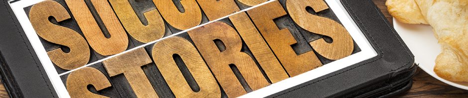 A tablet sits on a desk next to a cup of coffee and a croissant. Upon the tablet is a leather cover with the words Success Stories printed on it in lettering that looks like it is made out of stained wood.