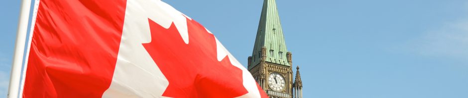 A large Canadian flag with a red maple leaf in the center waves prominently in the foreground against a clear blue sky. Behind it stands the iconic Peace Tower, part of Canada’s Parliament Buildings in Ottawa, with its green copper roof and a visible clock face. Atop the tower, a smaller Canadian flag is also flying, reinforcing the national symbolism of the scene.